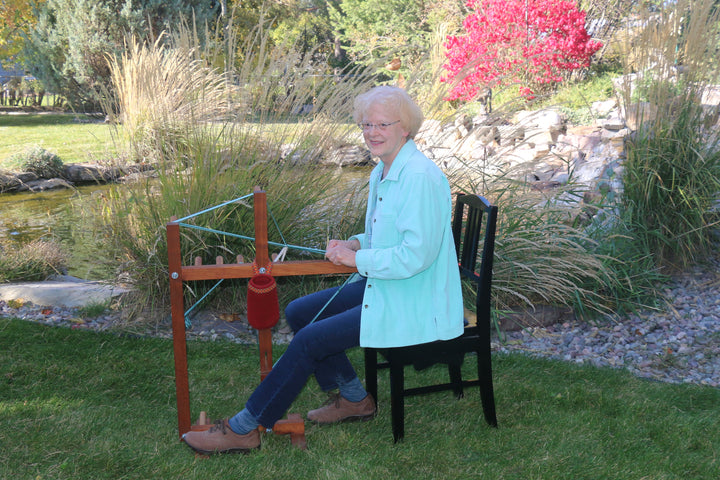 An older woman with short blond hair and glasses sits outdoors by a pond, spinning yarn on a wheel for Nordic projects, as featured in The Bandweaver's Project Book from Schiffer Publishing, Ltd.