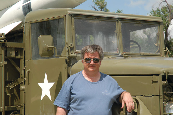 A man in sunglasses and a blue t-shirt stands by an olive green military truck with a missile, evoking the iconic Sd.Kfz. vehicles featured in Schiffer Publishing, Ltd.'s German Light Armored Cars of WWII.