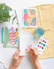 With Schiffer Publishing, Ltd.’s Watercolor the Easy Way Paint-Your-Own Watercolor Cards Garden kit, two finished cards—one floral, one with snakes—plus a pen and envelope sit on a yellow patterned cloth beside a glass of water and a plant.