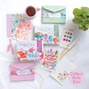 A flat lay of Schiffer Publishing, Ltd.’s Watercolor the Easy Way Paint-Your-Own Watercolor Cards Garden kit, featuring floral and mushroom cards, tea, paintbrushes, pencils, a palette, envelopes, and a “Collect Both Kits!” label on a light surface.