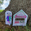 A hand-drawn paper cutout of a child in winter clothes stands beside a vibrant hot cocoa stand, both set against a tree trunk outdoors—evoking the cozy charm of the "Christmas Market Crew" coloring book by Schiffer Publishing, Ltd.