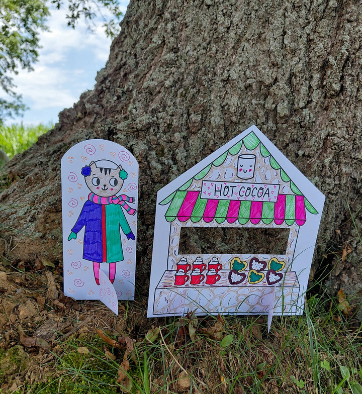 A hand-drawn paper cutout of a child in winter clothes stands beside a vibrant hot cocoa stand, both set against a tree trunk outdoors—evoking the cozy charm of the "Christmas Market Crew" coloring book by Schiffer Publishing, Ltd.