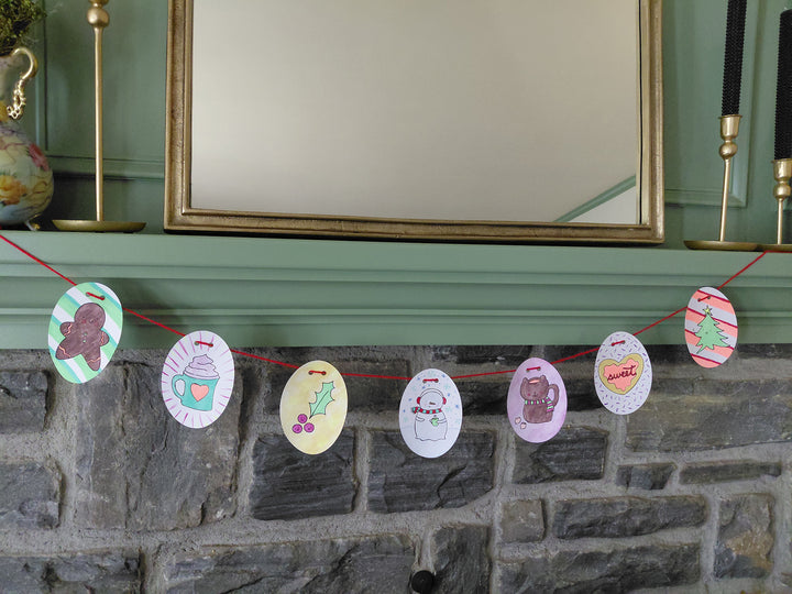 A string of oval-shaped paper ornaments from the "Christmas Market Crew" by Schiffer Publishing, Ltd., inspired by paper craft coloring book designs and adorned with festive holiday art, hangs on a red string across a gray stone fireplace beneath a green mantel and large mirror.
