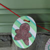 A paper ornament from Schiffer Publishing, Ltd.’s Christmas Market Crew features a smiling gingerbread person, strung on red yarn. Reminiscent of coloring book designs, the ornament displays green and white stripes in the background and hangs in front of a stone wall and a green shelf.