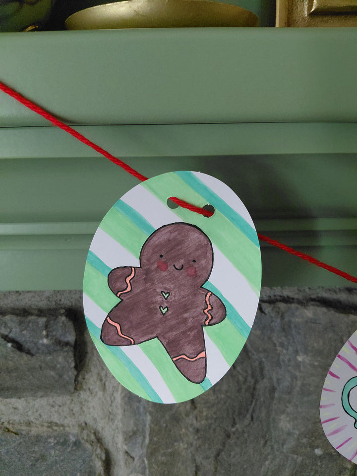 A paper ornament from Schiffer Publishing, Ltd.’s Christmas Market Crew features a smiling gingerbread person, strung on red yarn. Reminiscent of coloring book designs, the ornament displays green and white stripes in the background and hangs in front of a stone wall and a green shelf.