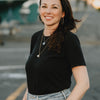 A woman with long brown hair in a black t-shirt and light blue jeans smiles while standing outdoors, holding the "Under the Sea Besties" coloring book from Schiffer Publishing, Ltd.