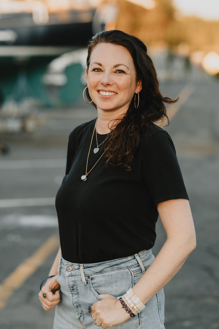 A woman with long brown hair in a black t-shirt and light blue jeans smiles while standing outdoors, holding the "Under the Sea Besties" coloring book from Schiffer Publishing, Ltd.