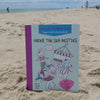 The “Under the Sea Besties” paper craft coloring book by Schiffer Publishing, Ltd. stands on a sandy beach with the ocean and people in the background. The cover shows cute aquatic characters and the subtitle "Color & Punch Fun.