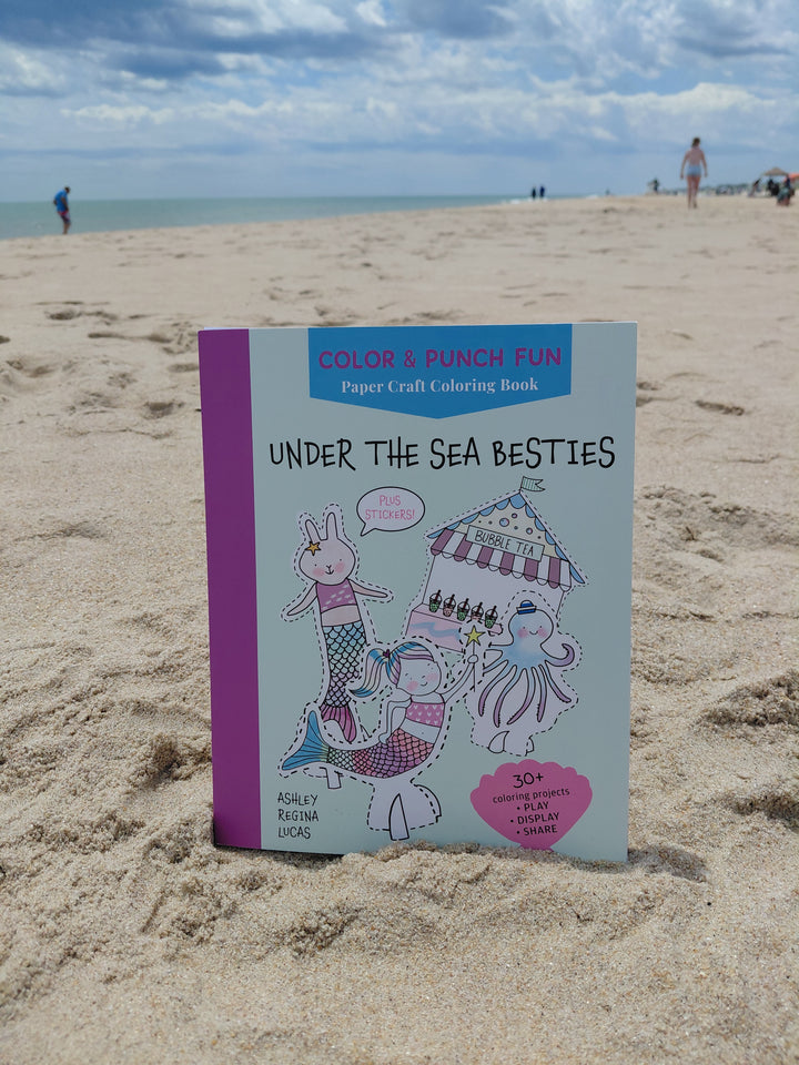 The “Under the Sea Besties” paper craft coloring book by Schiffer Publishing, Ltd. stands on a sandy beach with the ocean and people in the background. The cover shows cute aquatic characters and the subtitle "Color & Punch Fun.