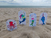 Under the Sea Besties by Schiffer Publishing, Ltd. features vibrant aquatic characters—octopus, mermaid, bunny-mermaid, and narwhal—on a sandy beach with an ocean backdrop, perfect for any ocean-themed coloring book.