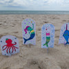 Under the Sea Besties by Schiffer Publishing, Ltd. features vibrant aquatic characters—octopus, mermaid, bunny-mermaid, and narwhal—on a sandy beach with an ocean backdrop, perfect for any ocean-themed coloring book.