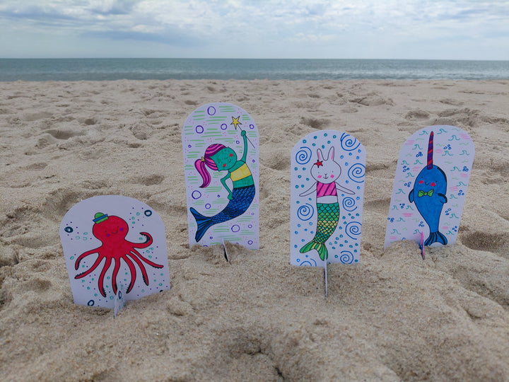 Under the Sea Besties by Schiffer Publishing, Ltd. features vibrant aquatic characters—octopus, mermaid, bunny-mermaid, and narwhal—on a sandy beach with an ocean backdrop, perfect for any ocean-themed coloring book.