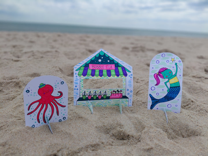Under the Sea Besties by Schiffer Publishing, Ltd. features three vibrant paper cutouts—a hat-wearing octopus, bubble tea stand, and smiling mermaid—decorating a sandy beach with an ocean-themed coloring book style.