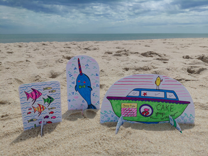 Colorful hand-drawn paper cutouts from Schiffer Publishing, Ltd.'s Under the Sea Besties—a fish, narwhal, and a Cake Pop Wagon boat—stand upright in the sand on a beach with the ocean and cloudy sky in the background.