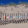 Under the Sea Besties" by Schiffer Publishing, Ltd. features colorful circular paper art inspired by ocean scenes, displayed on a wooden sand dune fence with tall grass, smooth sunlit sand, and a deep blue sky in the background.