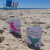 Two vibrant "Under the Sea Besties" cards from Schiffer Publishing, Ltd. stand on a sandy beach—one with a loving seal, the other with a mermaid tail—perfect inspiration for ocean-themed coloring fans. Two chairs face the clear blue sky nearby.