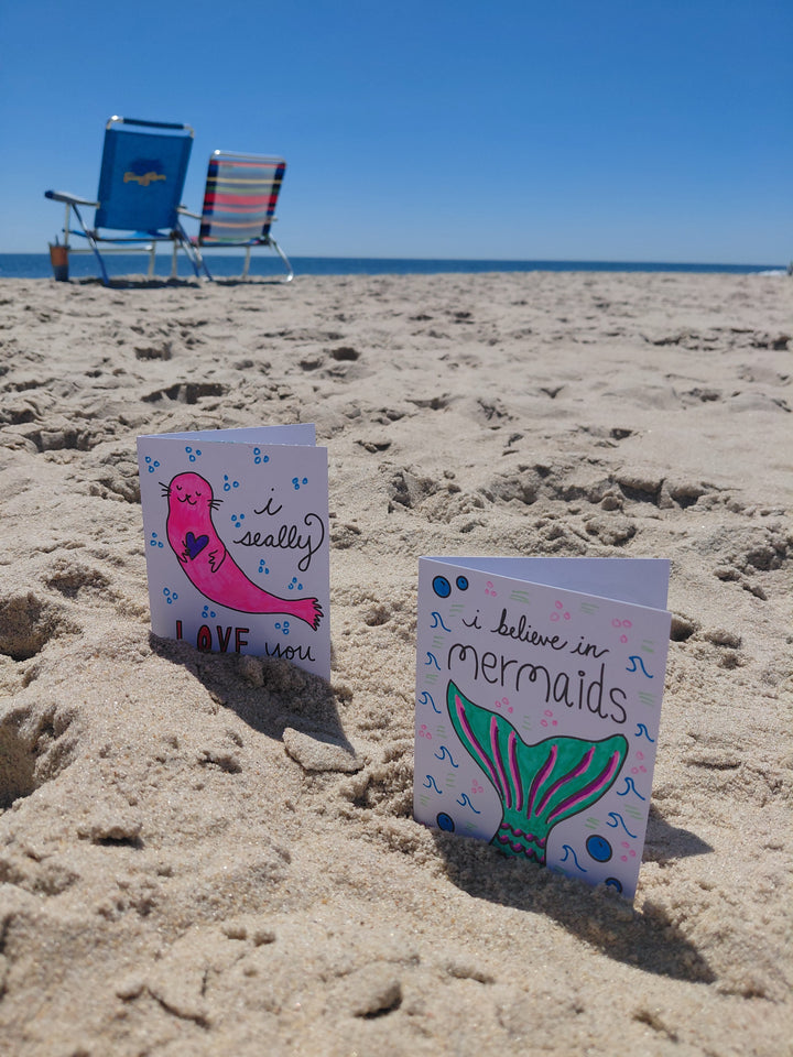Two vibrant "Under the Sea Besties" cards from Schiffer Publishing, Ltd. stand on a sandy beach—one with a loving seal, the other with a mermaid tail—perfect inspiration for ocean-themed coloring fans. Two chairs face the clear blue sky nearby.