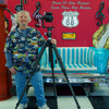 A smiling man in a Route 66 shirt stands by a camera beside a teal and white bench. Behind him are Route 66 signs, “Rock ’n’ Roll is here to stay” decor, and Yellowstone National Park art from Schiffer Publishing, Ltd.
