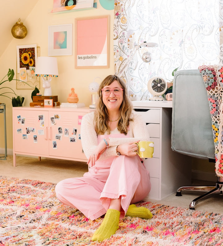A woman with long hair and glasses, dressed in pink overalls and yellow socks, sits on a colorful rug in a pastel-themed room, smiling at the camera while holding Schiffer Publishing’s Good Vibes Art Club Learn to Draw book.