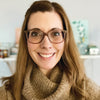 A smiling woman with long light brown hair and glasses holds "The Very Cozy Coloring Book" by Schiffer Publishing, Ltd. She wears a tan turtleneck sweater, surrounded by warm-toned decor in a softly lit, inviting room.