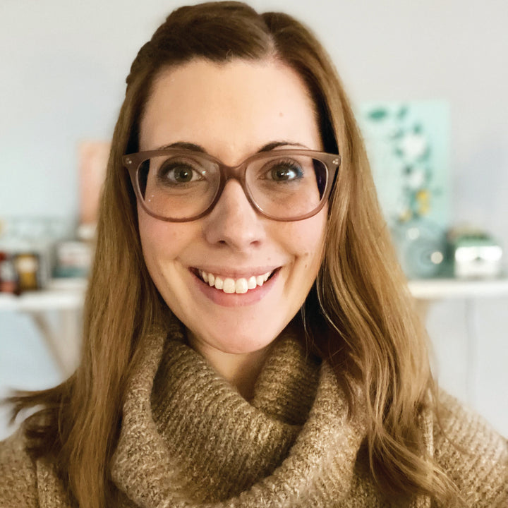 A smiling woman with long light brown hair and glasses holds "The Very Cozy Coloring Book" by Schiffer Publishing, Ltd. She wears a tan turtleneck sweater, surrounded by warm-toned decor in a softly lit, inviting room.