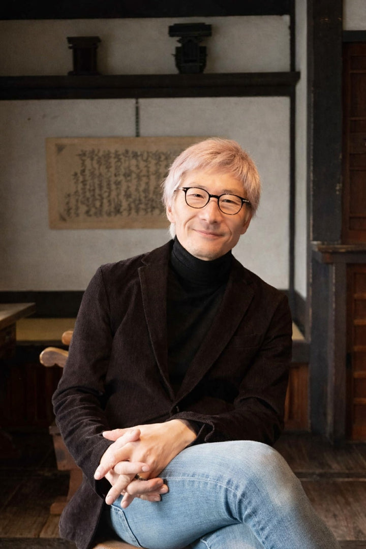 A man with light hair and glasses smiles in a traditional Japanese-style room with wooden walls made using joinery, next to "Traditional Japanese Woodworking" by Schiffer Publishing, Ltd., wearing a black turtleneck, brown jacket, and jeans.