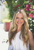 A woman with long blonde hair and a white sleeveless top smiles outdoors near pink flowers, holding Schiffer Publishing, Ltd.'s Watercolor the Easy Way Paint-Your-Own Watercolor Cards Autumn kit in soft sunlight.