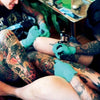 A tattoo artist in green gloves inks a client’s tattooed leg, both adorned with multiple tattoos. This vibrant scene, set in a bright studio, is captured in "AOI Ink Nymphs Los Angeles" by Schiffer Publishing, Ltd.
