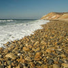 Experience the beauty of Martha’s Vineyard Wide by Schiffer Publishing, Ltd.—smooth stones along a rocky shore lead to ocean waves and a clear sky, with distant cliffs reminiscent of iconic coastal panoramas.