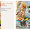 An outdoor spread of Mediterranean dishes features salads, grilled vegetables, flatbread, clams, and juice. Someone holds salad as the left shows "Grill Skills: Professional Tips for the Perfect Barbeque" by Schiffer Publishing, Ltd. with grilling tips.