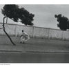 A person in flowing clothing walks beside bent trees and a tall white wall under clouds, echoing scenes from "Timeless" by Schiffer Publishing, Ltd.; another person crouches by a tree at the right edge.