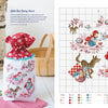 A drawstring bag with Véronique Enginger’s Little Red Riding Hood cross stitch from Schiffer Publishing, Ltd.’s "Fables & Fairy Tales to Cross Stitch" sits on a table beside flowers, mushrooms, and the matching pattern chart.