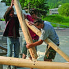 Workers assemble large wooden logs outdoors using sustainable techniques from "Natural Building" by Schiffer Publishing, Ltd. Trees and a building are in the background, with tools and wood shavings scattered nearby.
