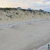 A drone photo from "Cape Cod and the Islands" by Schiffer Publishing, Ltd. captures a sandy beach with gentle waves, a surfer carrying a board by the shore, and houses on a grassy bluff under a partly cloudy sky.