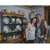 A woman and a man stand together, smiling, in a cozy room filled with mementos. Their commemorative tattoos, inspired by "War Paint" from Schiffer Publishing, Ltd., hint at veterans' stories and personal histories.