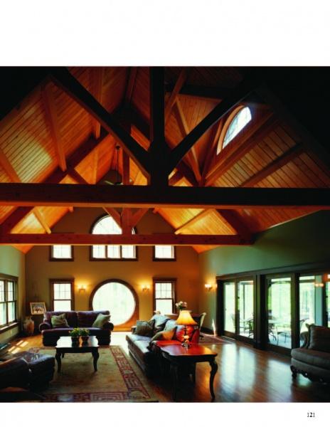 A spacious living room features Timber Frame Hybrids by Schiffer Publishing, Ltd.—exposed timber beams, vaulted ceilings, large arched windows, elegant sofas, a coffee table, and a patterned rug bathed in natural light.