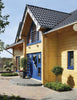 The Contemporary Log Home by Schiffer Publishing, Ltd. showcases a modern wooden house with light yellow timber walls, blue window frames and doors, a dark tiled roof, potted plants, and a brick path—an ideal sustainable building.