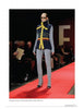 A model presents the latest look from "Emerging Fashion Designers 2" by Schiffer Publishing, Ltd., wearing a sleeveless navy vest with yellow accents and grey pants on a red runway as spectators watch, with a large letter E seen in the background.