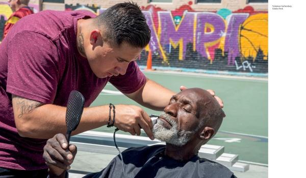A barber trims an elderly man's beard outdoors as he relaxes in a black cape. Behind them, colorful graffiti and a basketball court embody the spirit of Design with Love by Schiffer Publishing, Ltd., highlighting vibrant community development.