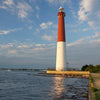 Barnegat Lighthouse Perspectives by Schiffer Publishing, Ltd. features the iconic red and white Barnegat Lighthouse on Long Beach Island, set beside calm water with sunlit reflections, rocky shoreline, fence, and a partly cloudy sky.