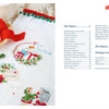 A festive cloth with Christmas embroidery and a red ribbon rests next to "The Magic of Christmas to Cross Stitch" by Schiffer Publishing, Ltd., opened to a table of contents for holiday cross-stitch projects.