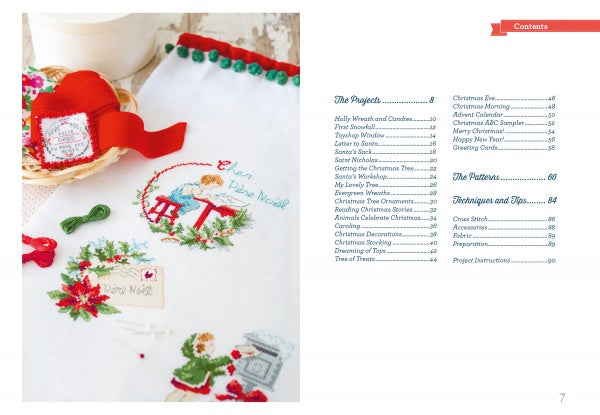 A festive cloth with Christmas embroidery and a red ribbon rests next to "The Magic of Christmas to Cross Stitch" by Schiffer Publishing, Ltd., opened to a table of contents for holiday cross-stitch projects.