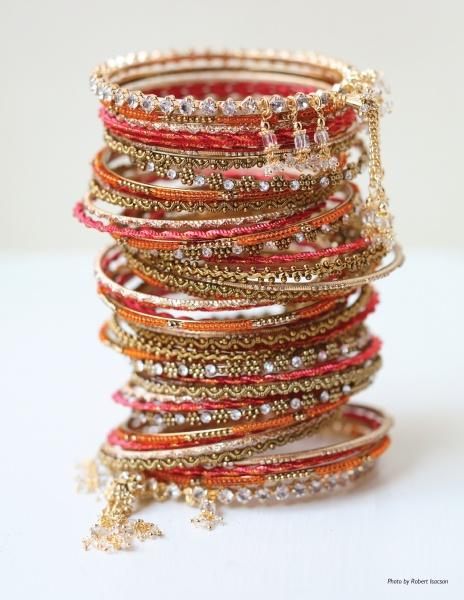 A stack of vibrant, ornate bangles with gold, silver, and red accents—ideal for Indian Weddings by Schiffer Publishing, Ltd.—features intricate patterns and dangling gold chains, elegantly displayed on a white surface.