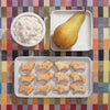 A bowl of cottage cheese, a whole pear, and twelve animal-shaped treats from "Baking for Dogs" by Schiffer Publishing, Ltd. are displayed on a colorful checkered tablecloth.