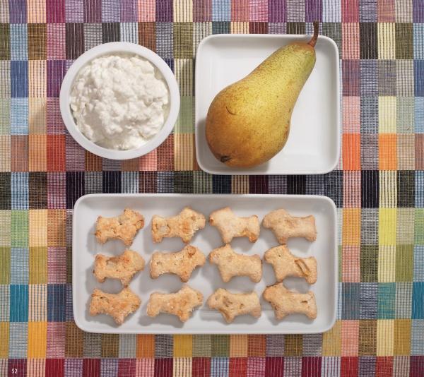 A bowl of cottage cheese, a whole pear, and twelve animal-shaped treats from "Baking for Dogs" by Schiffer Publishing, Ltd. are displayed on a colorful checkered tablecloth.
