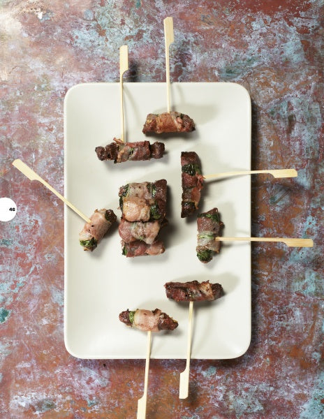 A rectangular white plate holds seven bacon-wrapped appetizers on wooden picks, inspired by classic recipes from "Pure BBQ!" by Schiffer Publishing, Ltd., arranged on a textured, multicolored surface.