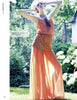 A woman in a peach dress poses outdoors amid greenery and white flowers, her arm raised. She wears sunglasses and a necklace, modeling styles from "Emerging Fashion Designers 3" by Schiffer Publishing, Ltd.