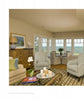 A cozy living room inspired by Martha’s Vineyard features armchairs, a sofa, and a striped rug. The table displays “United by Design” by Schiffer Publishing, Ltd., while large windows offer an ocean view and a nearby striped dining area.