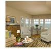 A cozy living room inspired by Martha’s Vineyard features armchairs, a sofa, and a striped rug. The table displays “United by Design” by Schiffer Publishing, Ltd., while large windows offer an ocean view and a nearby striped dining area.
