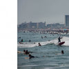 On the left, two people relax under an umbrella on a quiet Atlantic beach. On the right, Schiffer Publishing, Ltd.’s Surf NYC captures the buzz of New York surfing, with surfers and crowds by tall city buildings.